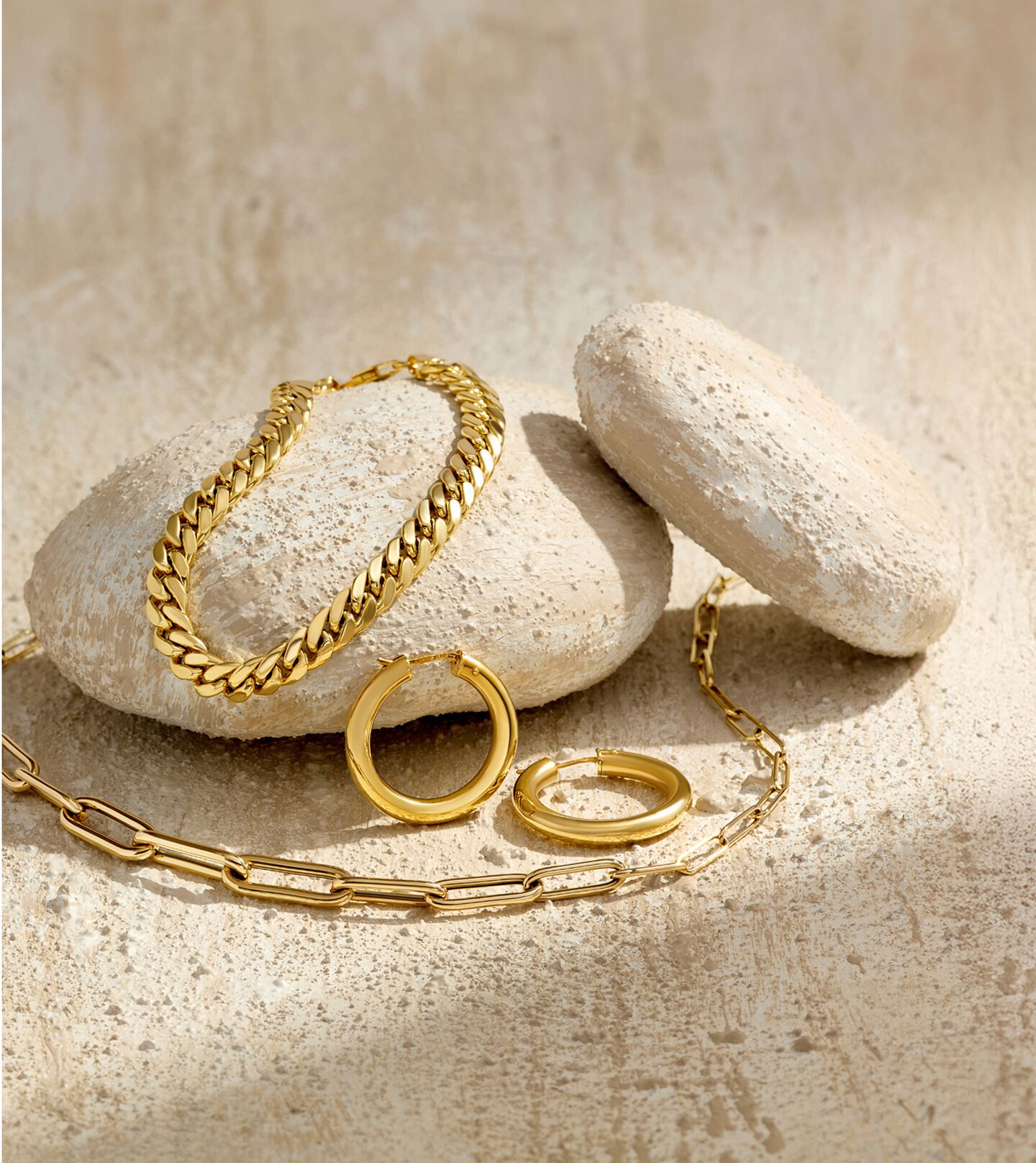 14k yellow gold bracelet, hoop earrings and necklace rest on white rocks that cast shadows over the surface of a white stone table. 