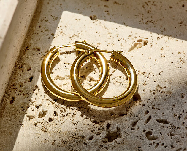 Gold Italian crafted hoop earrings overlap on a flat stone surface while the shining hoops reflect light. 