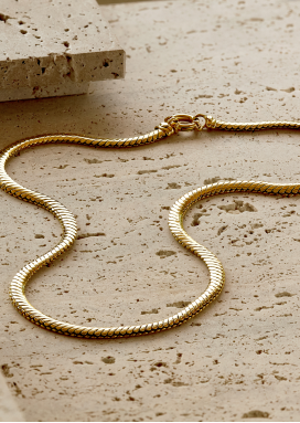 A yellow gold necklace with flowing links and a lobster clasp rests on a surface of flat porous stone. 