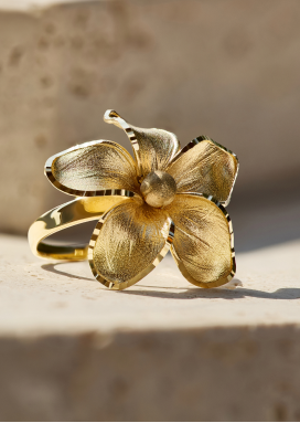A floral ring with petals of 14k yellow gold casts shadows on a textured off-white background.